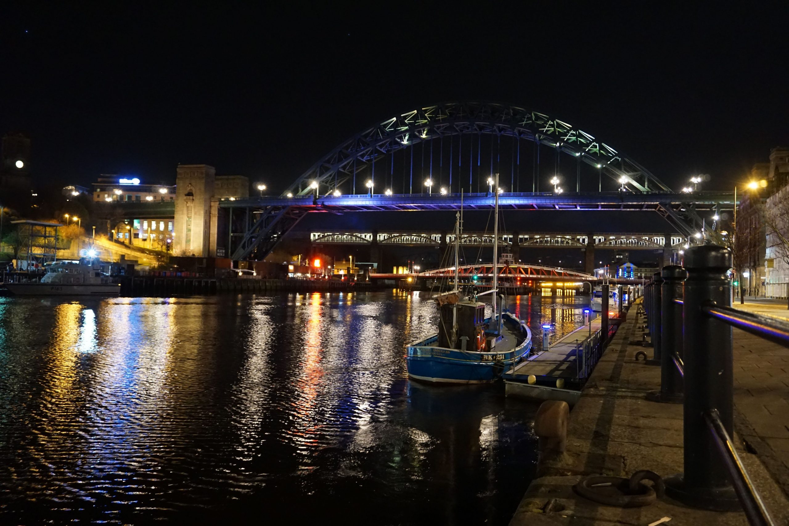 A small boat is moored in the River Tyne at night, the background has the Tyne Bridge. Lights are reflected in the water.